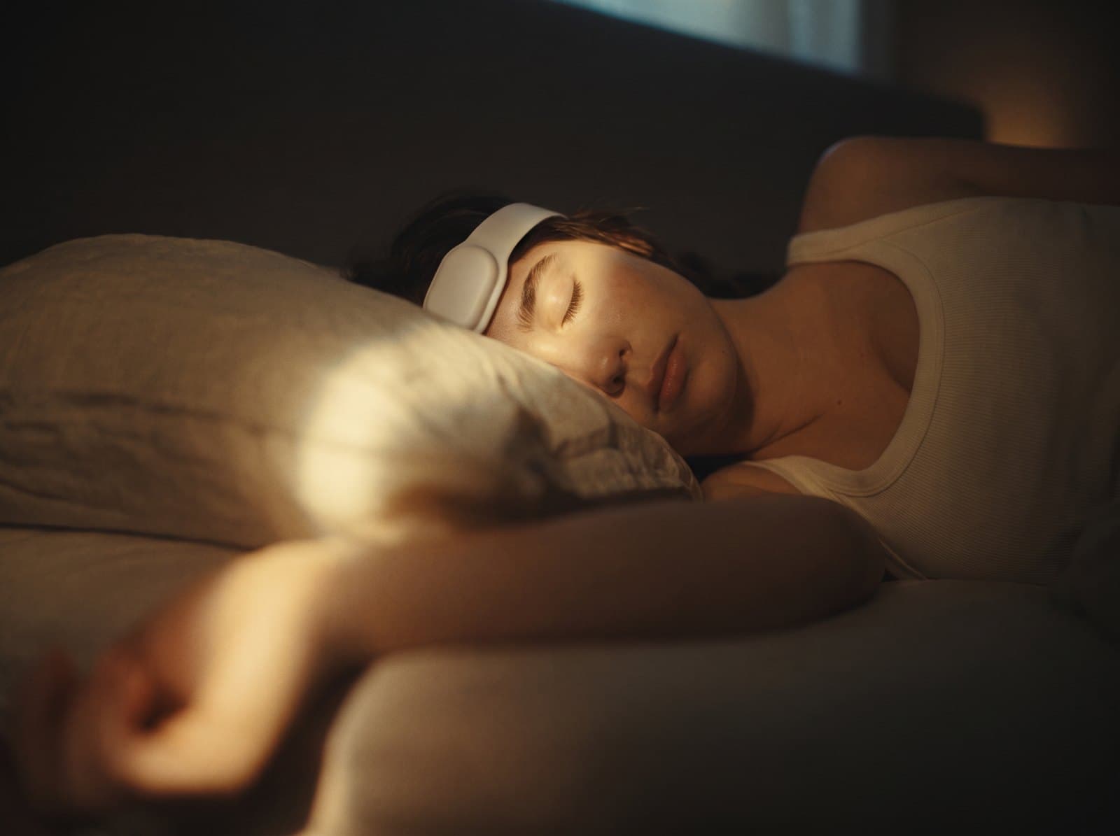 A woman asleep in a dark bedroom, the wearable on her forehead lit by a bedside lamp.
