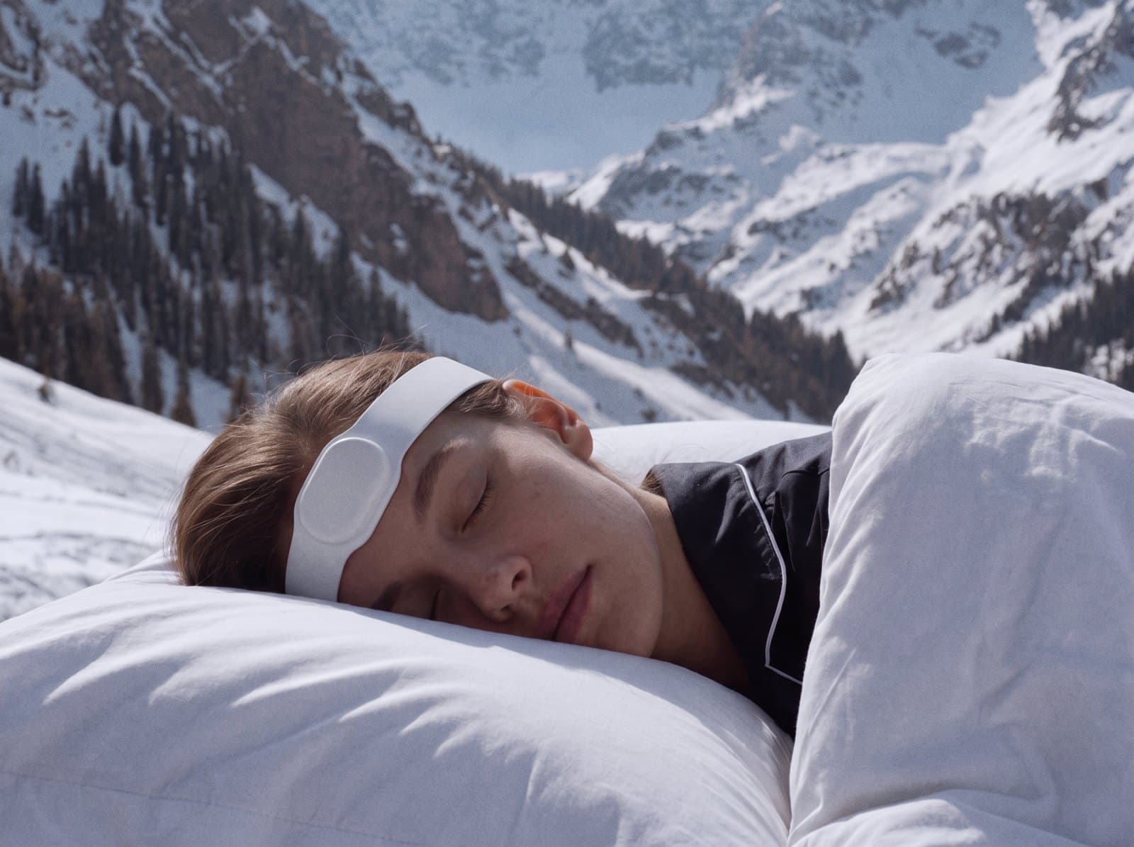 A woman asleep in white bedding under snowy mountains, wearing the Paprika wearable.
